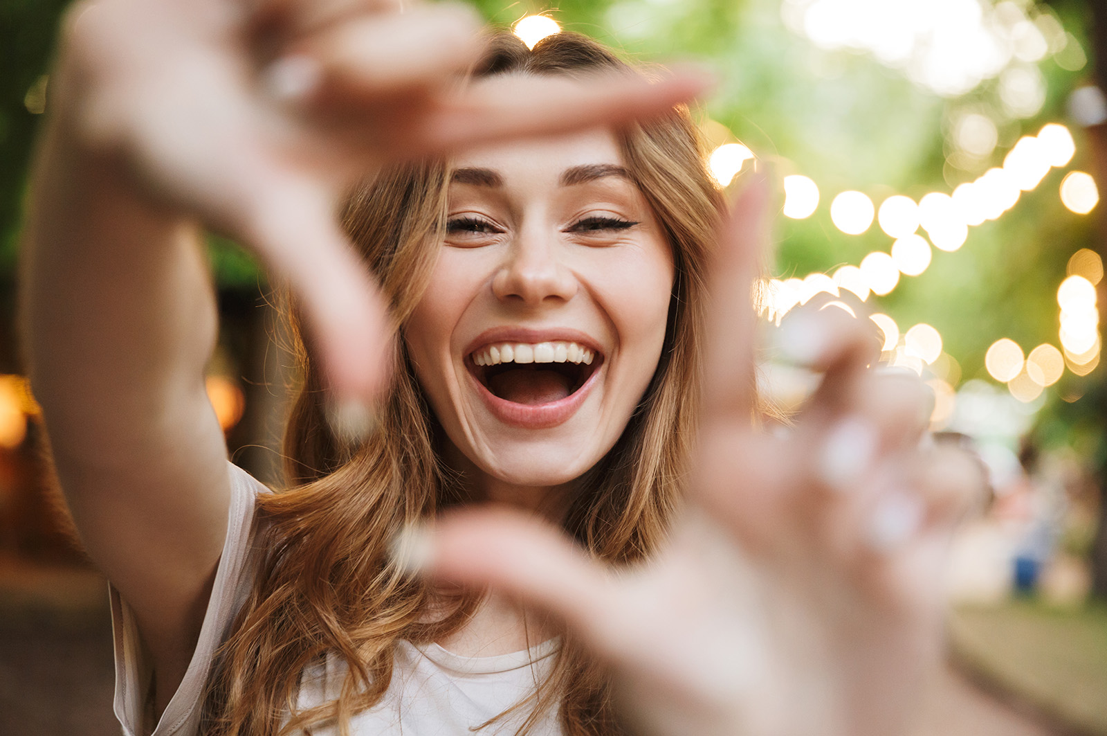 A young woman with her hands up near her face, smiling broadly at the camera while standing outdoors during daylight hours.