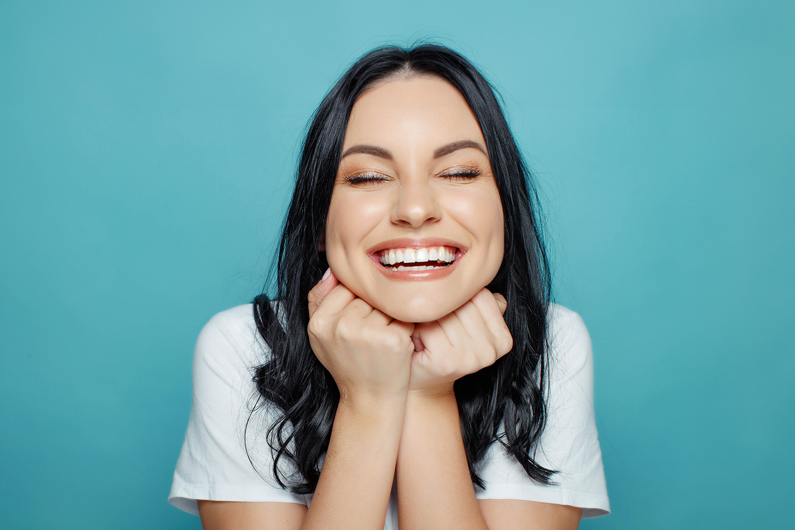 A woman with dark hair is smiling at the camera while holding her hands together near her face.