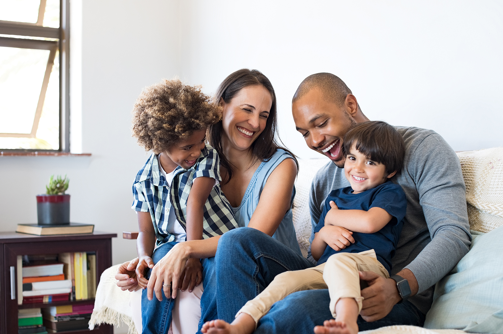 The image shows a family sitting on a couch, with a man holding a child and two other individuals looking at him. They appear to be engaged in a moment of shared attention, possibly during a casual indoor setting.