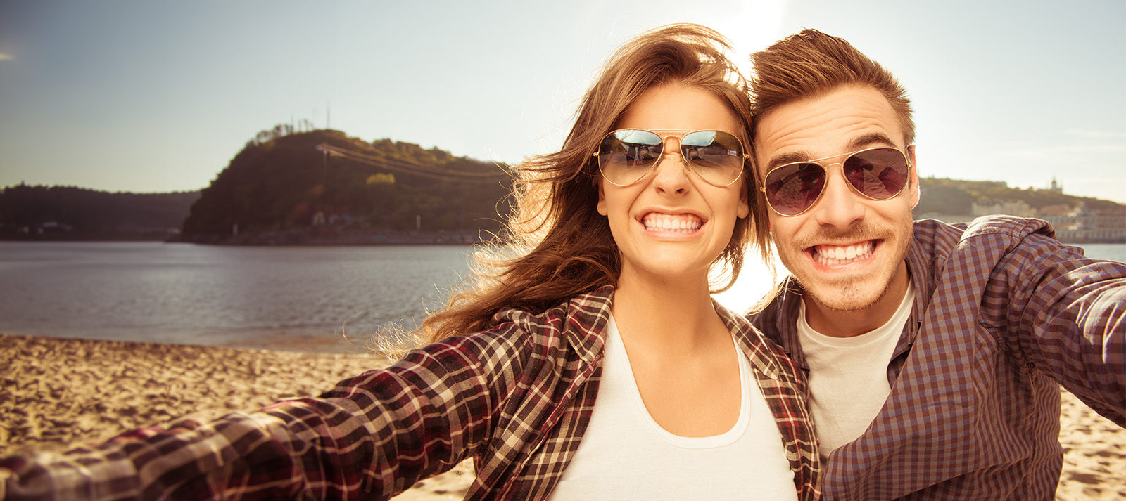 A young couple posing for a selfie on a sandy beach with clear skies, both smiling broadly.