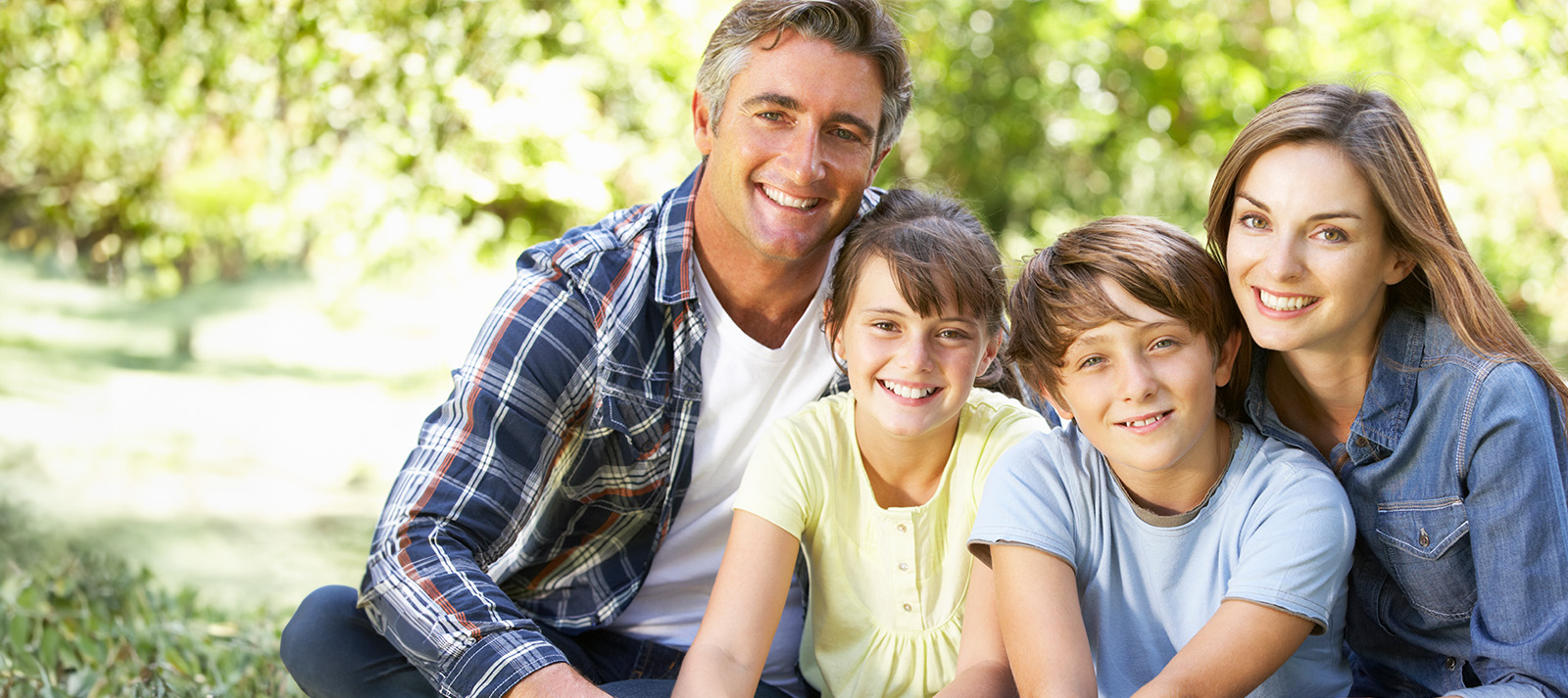 The image shows a family of four people, including two adults and two children, sitting outdoors on grass with trees in the background.