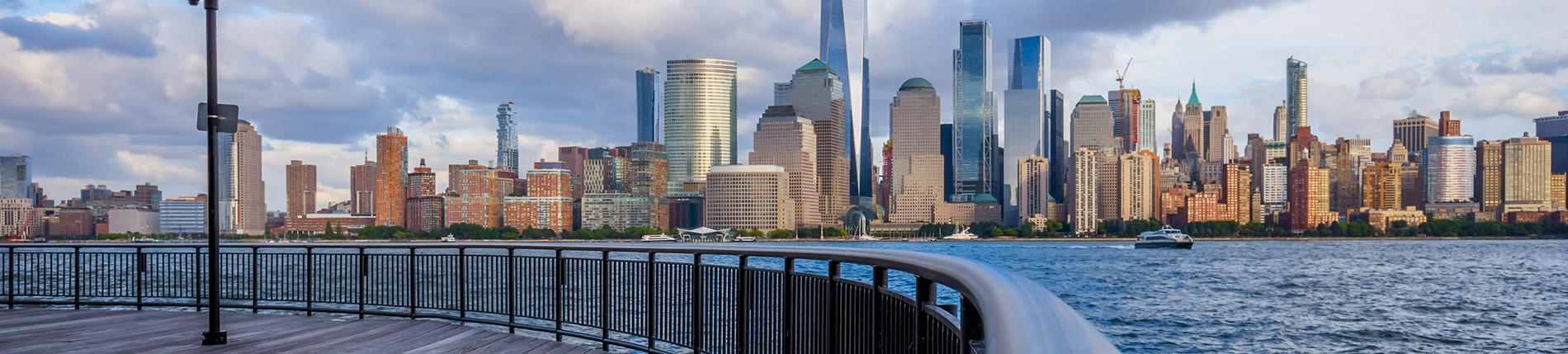 The image shows a city skyline with a prominent skyscraper and a body of water in the foreground, viewed from an elevated perspective, possibly a bridge, during daylight hours under partly cloudy skies.