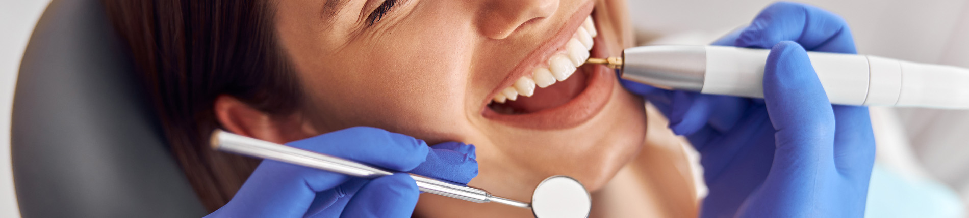 The image shows a person receiving dental treatment with a dental hygienist using a dental mirror and probe, while wearing blue gloves and a surgical mask.