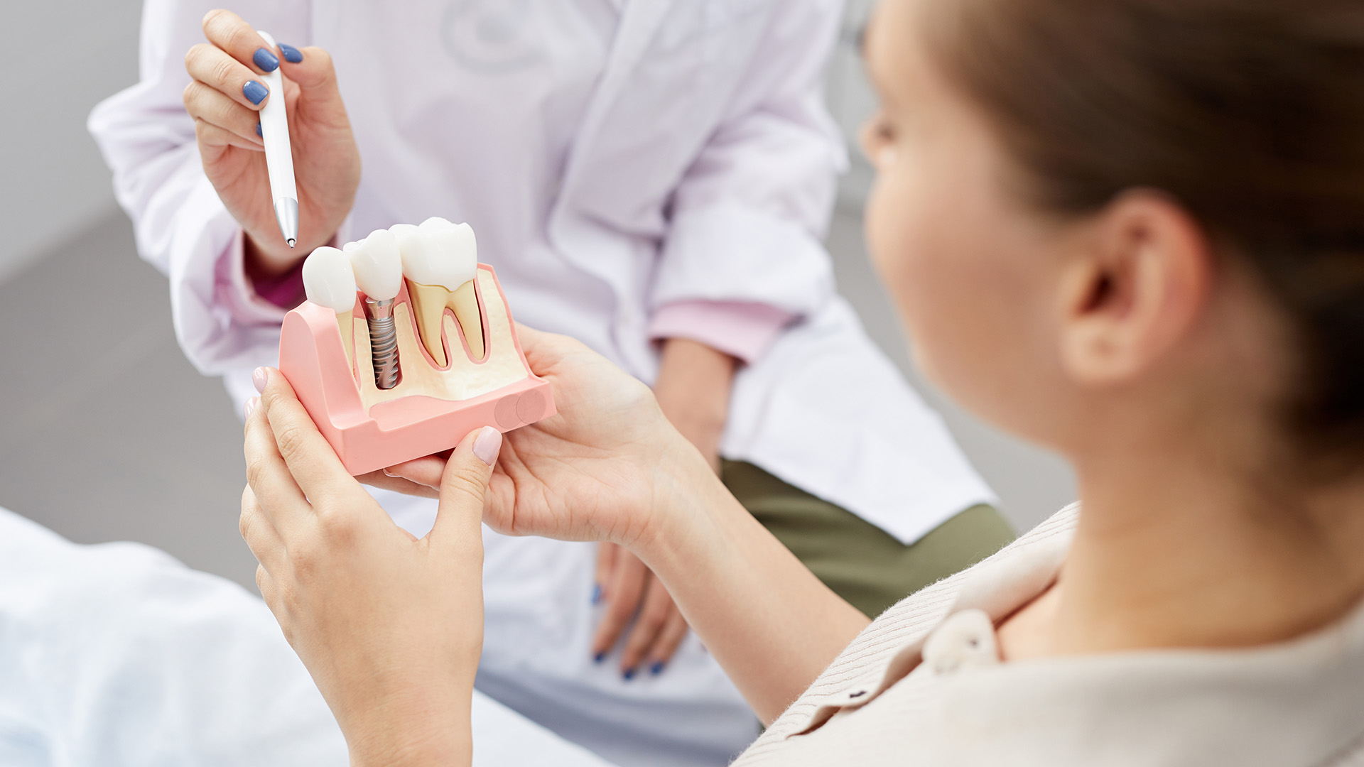 The image shows a person holding a pink model of a mouth with teeth and gums, while another individual appears to be observing or assisting the first.