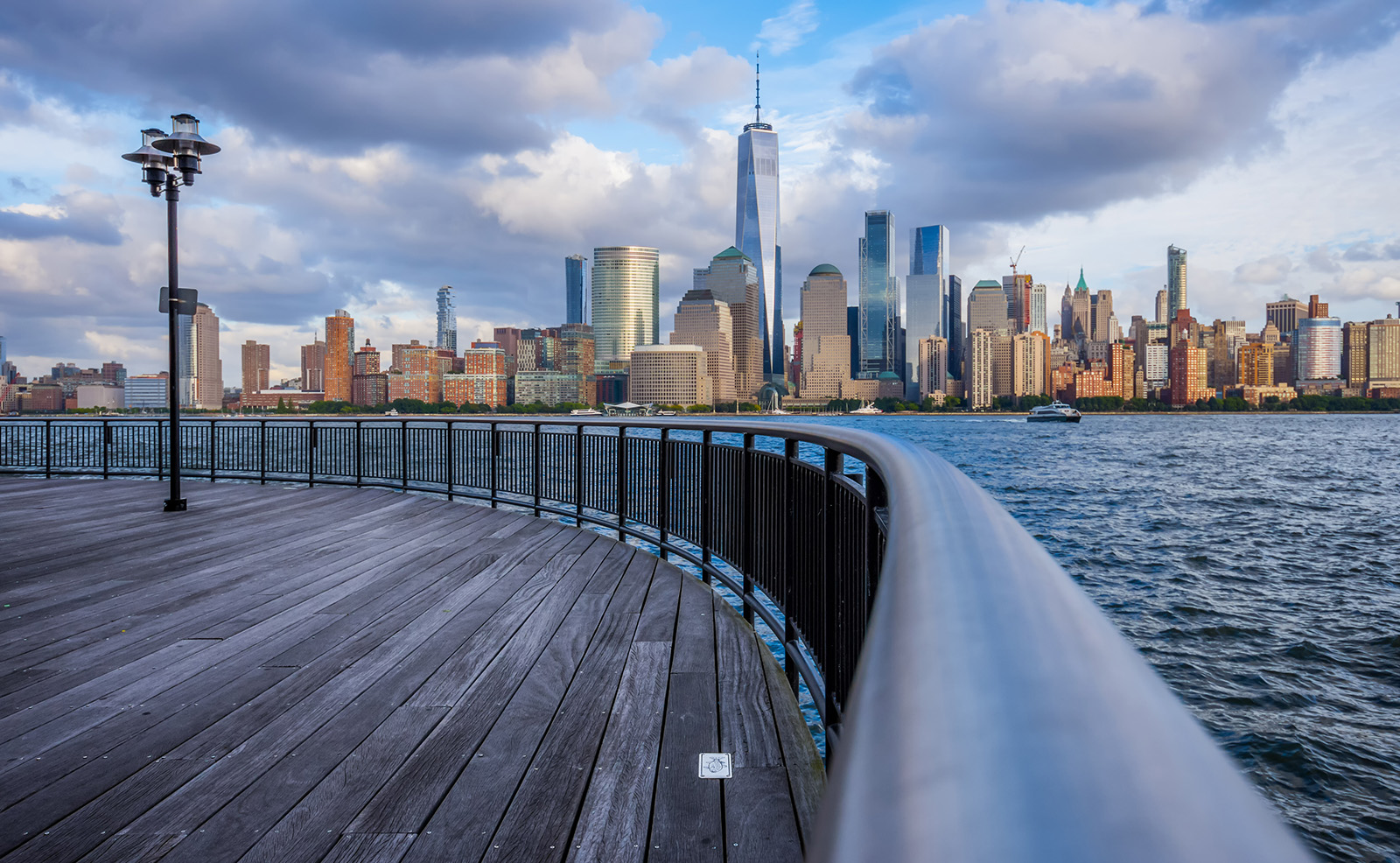 A photograph depicts a scenic view of a city skyline with the Manhattan skyline in the background, taken from a pier overlooking a river, with a clear sky above.