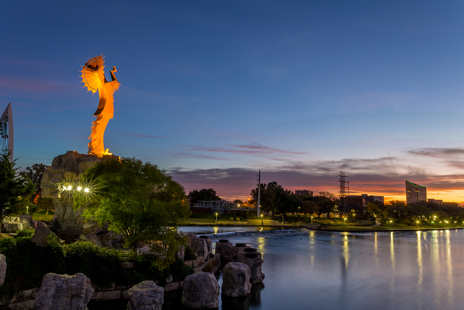 The image features a statue illuminated against a dusk sky, with a body of water reflecting the light, and a park setting with trees and a pier visible in the foreground.