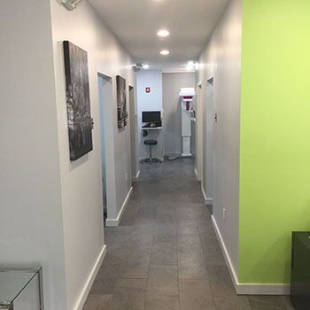 The image shows an interior hallway with a clean and minimalist design, featuring white walls, gray flooring, and a long hallway leading to a brightly colored room at the end.