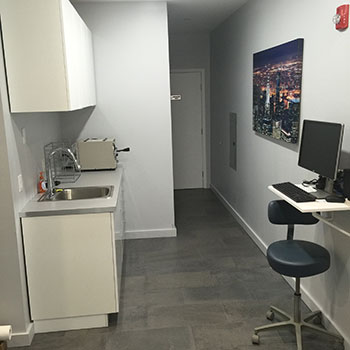 The image shows an interior space with walls painted white, featuring a kitchenette area equipped with a sink, countertop, and appliances, adjacent to a small desk with a computer monitor and keyboard, and a stool for seating. There s a window with a view of a cityscape, and the flooring appears to be a light-colored wood or laminate. The room is well-lit and appears clean and organized.