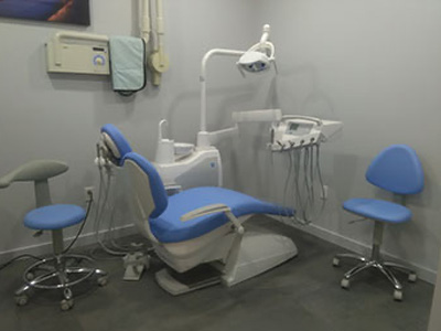 The image shows a dental office setup with a chair, a dental table, and equipment, including a dental chair, overhead lights, and various tools, all displayed against a wall with a blue chair for patient seating.