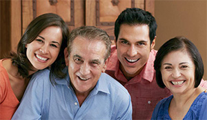 A family posing together with smiles, featuring an older man, woman, and younger couple, all dressed casually, set against a warm-toned background.