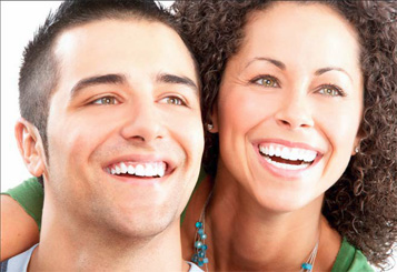 The image shows two individuals smiling at the camera  the person on the left appears to be male with short hair, while the one on the right seems female with long hair. They are close together, suggesting a relationship, and both are looking directly at the viewer with bright smiles.