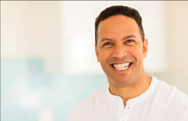 The image shows a man with a smile, wearing a white shirt, against a blurred background that appears to be an indoor setting.