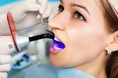 The image shows a woman receiving dental care with a dental drill and purple dye used for cavity detection, while wearing protective eyewear and gloves.