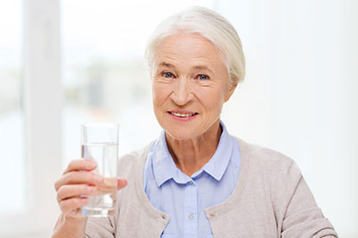 The image shows an elderly woman holding a glass of water while smiling at the camera.