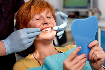 The image features a woman wearing a surgical mask, sitting in a dental chair with her mouth open, receiving dental care from a professional who appears to be adjusting her teeth using a dental mirror and instrument.