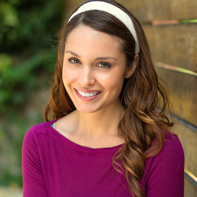 A young woman with long hair, wearing a purple top, stands against a wooden fence with a smile on her face.