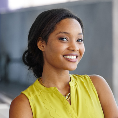 A young woman with a radiant smile, posing against a blurred background, wearing a yellow top and a necklace, with her hair pulled back, and she has a confident stance.