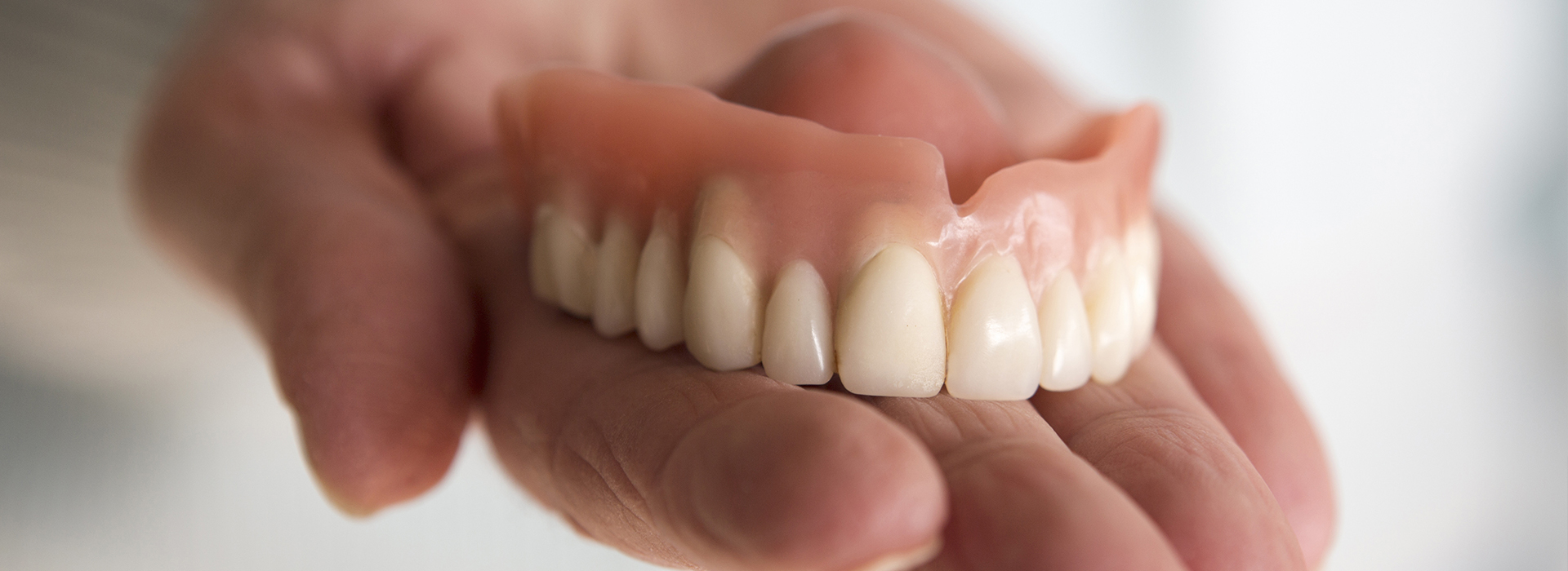 An adult hand holding a set of dentures against a light background.