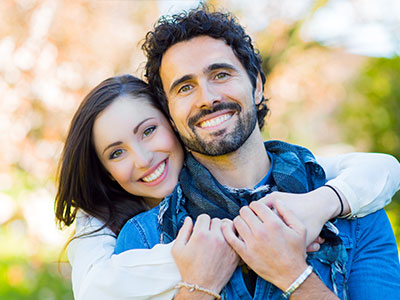 A man and woman are embracing each other in an outdoor setting, with the man wearing a scarf around his neck.