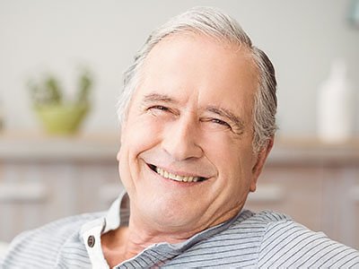 An elderly man with gray hair, smiling broadly at the camera while seated comfortably indoors.