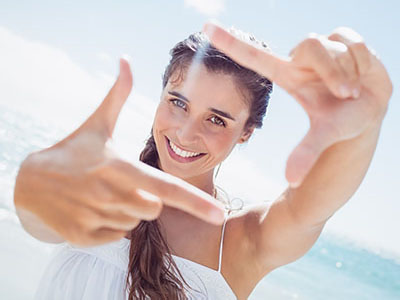 A woman with long hair is taking a selfie at the beach, holding up her hand to frame the photo.