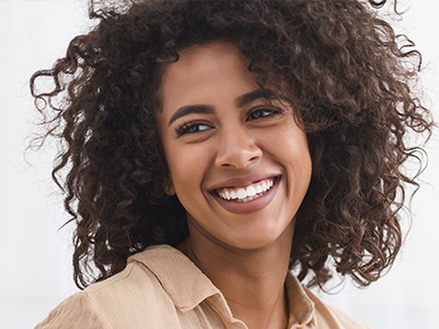 The image shows a smiling woman with curly hair, wearing a brown top, against a plain background.